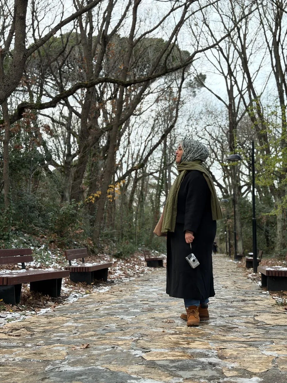 Kabátos, sállal és sapkával öltözött középkorú nő egy park sétányán áll, a kezében egy kis fényképezőgéppel.